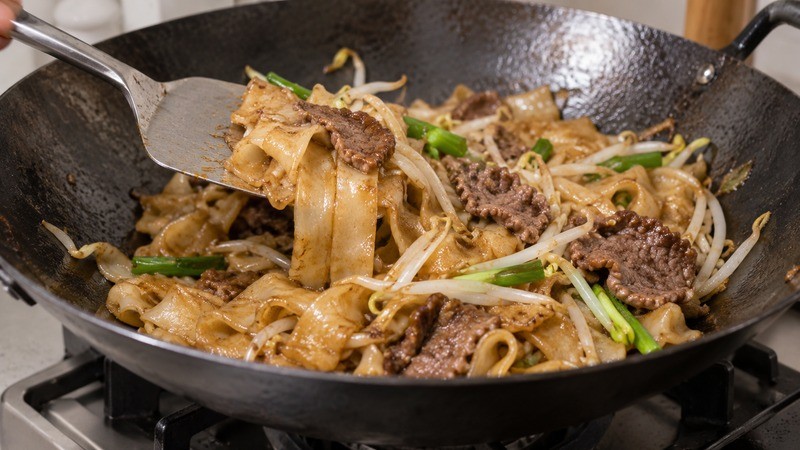 A spatula folding wide rice noodles with beef and thoroughly heated bean sprouts