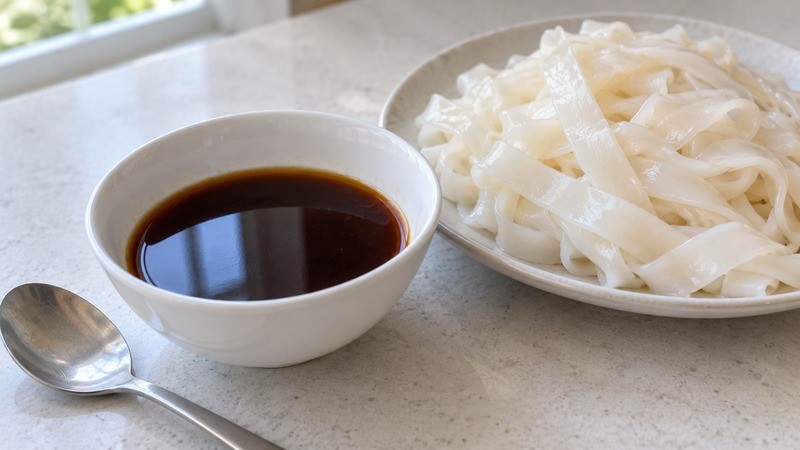 A small bowl of mixed thin brown seasoning sauce beside wide white rice noodles