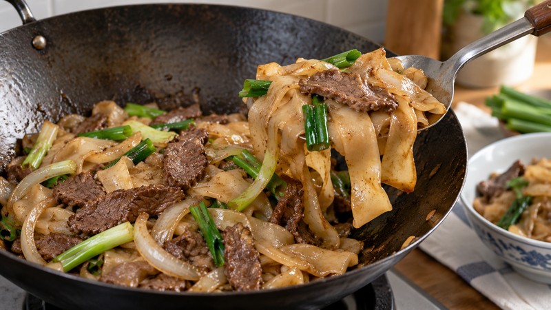 Broad spatula lifting wide rice noodles in a wok