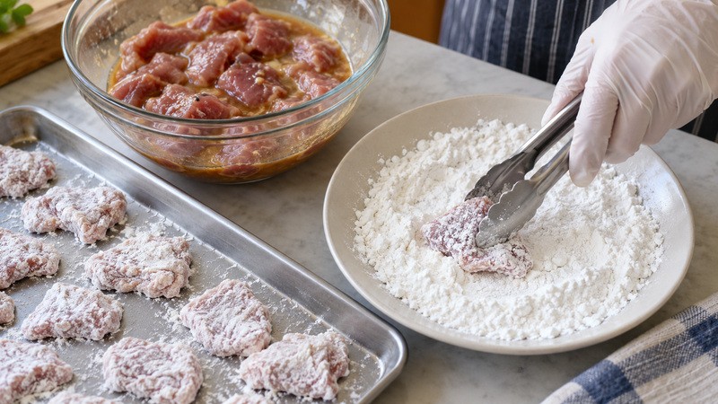 Thin pork shoulder pieces being individually coated in cornstarch beside marinade bowl