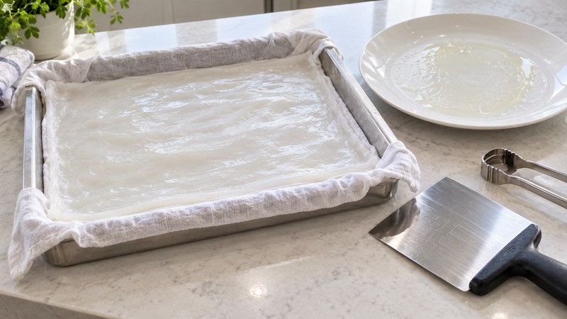 Thin rice sheet on a cotton-lined tray beside a scraper